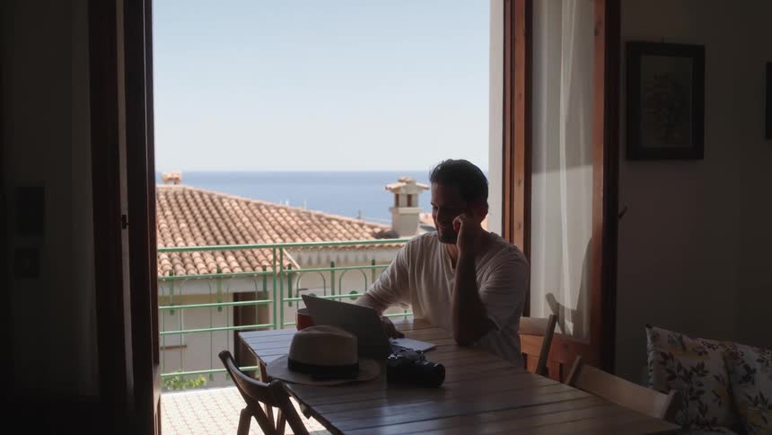 Man sitting at a table talking on the phone and using a laptop next to an open window showing the sea and a house on the same neighborhood during day time