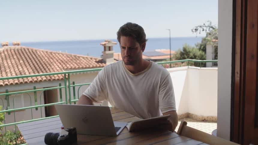A man is sitting at a table on a balcony with a laptop computer and writing in a book