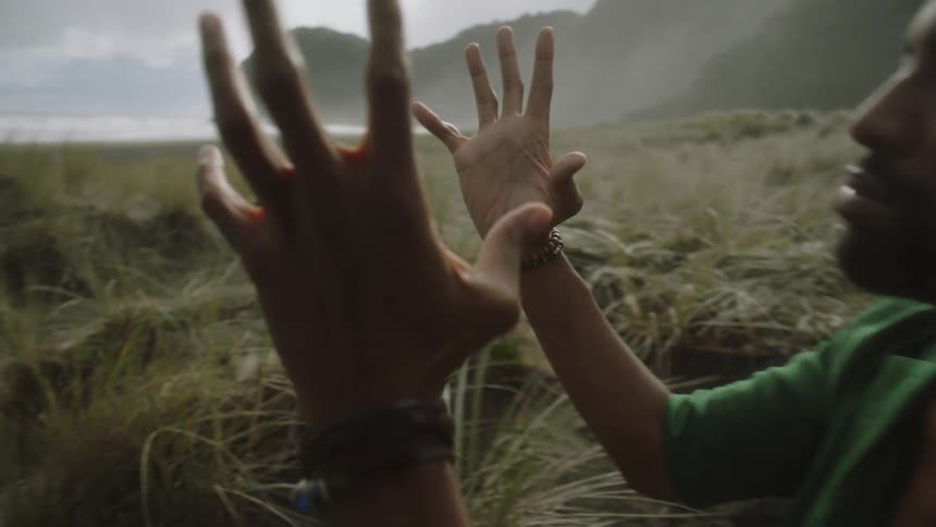 Man sitting in the grass next to the coast gestures mystically with his hands