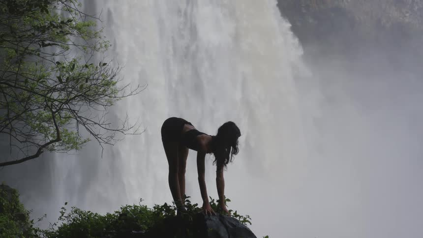 A woman on top of a rock practicing yoga in a natural environment with a waterfall in the background
