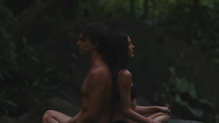 A man and a woman seated back to back in a yoga position in a natural environment with abundant vegetation