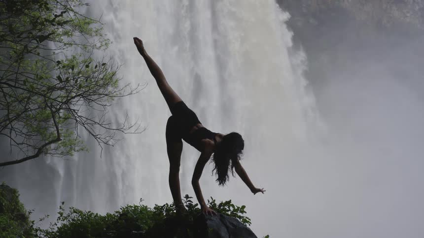 A woman doing yoga on a cliff overlooking a large waterfall during the day