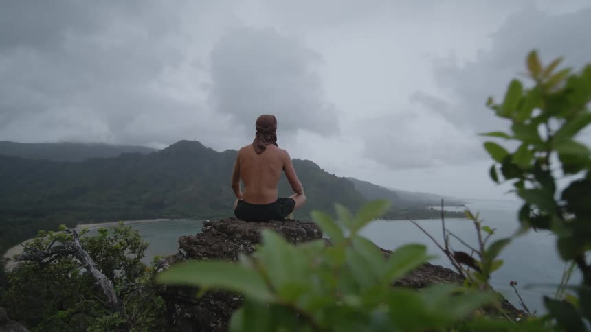 A man is sitting in a yoga pose on a rock cliff overlooking the sea on an overcast day