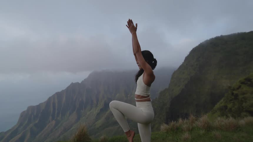 A young woman practicing yoga at the edge of a cliff with mountain view in sight as the sky is cloudy