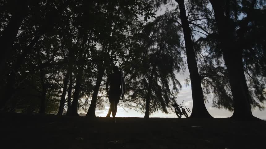 A man with a surfboard in his arms walks through a forest