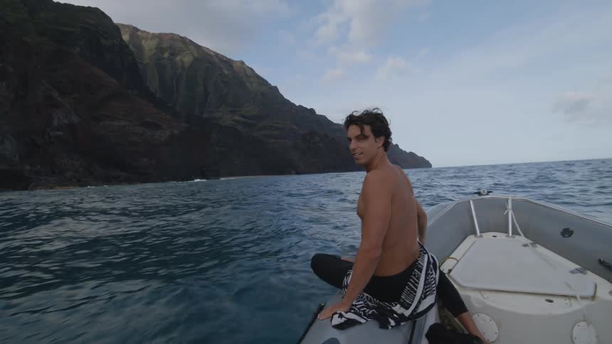 A man looks around at his surroundings while sitting on the edge of a motorboat as it travels along the coast during the day