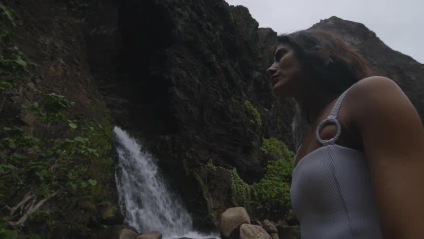 A woman sitting on a rock close to a stream near a water fall surrounded by rocks and green vegetation