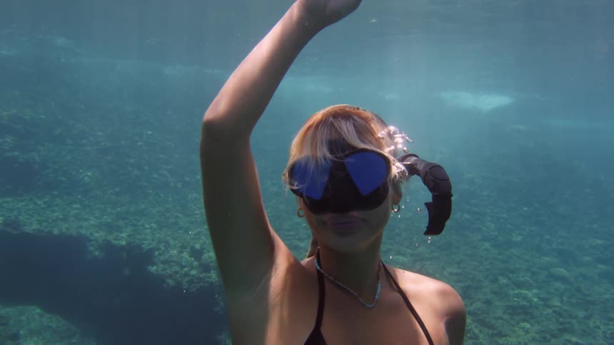 A woman wearing goggles and a snorkel while spinning around under water towards the surface