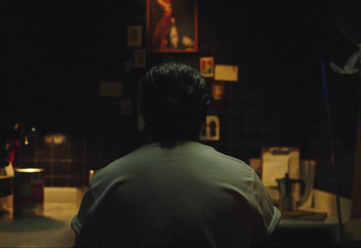 A man sits with his back to the camera in a room staring at pictures on a tiled wall