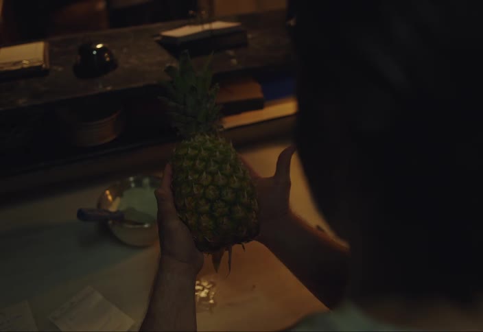 A person looking at a pineapple on their counter picking it up and then cutting it with a knife in a low lit room