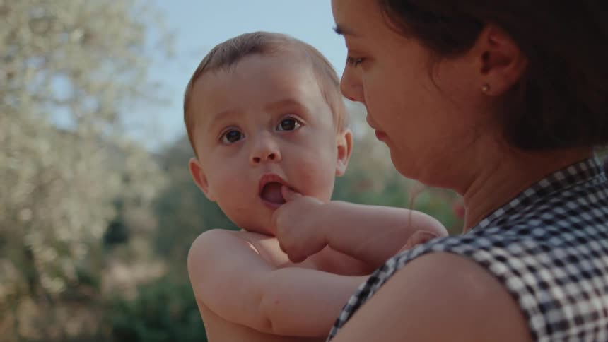 Camera moves towards a mother holding her shirtless baby in an outdoor setting with sunlight