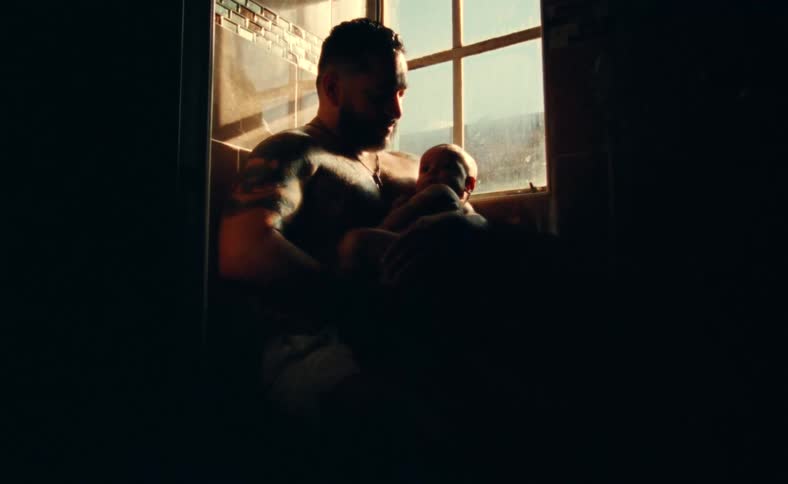 Strong muscular tattooed father sits with his newborn baby next to a window