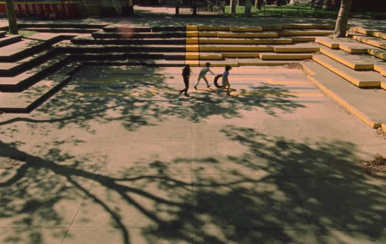 Three children run and roll a tire on the ground in a park next to stairs