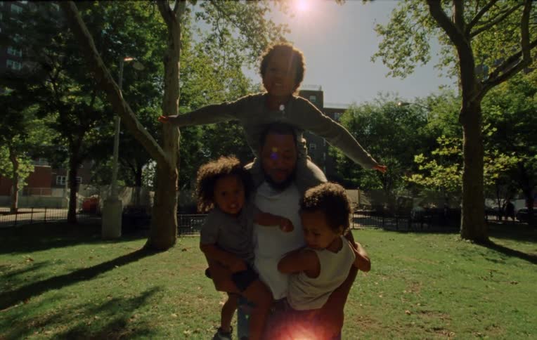 A father in a park holding three toddlers two under his arms with one of them slipping down his leg and another on his shoulder flapping his arms like a bird