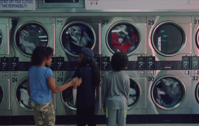 Two children with their backs turned looking at a washing machine from inside a laundry room when a girl arrives and starts talking to them