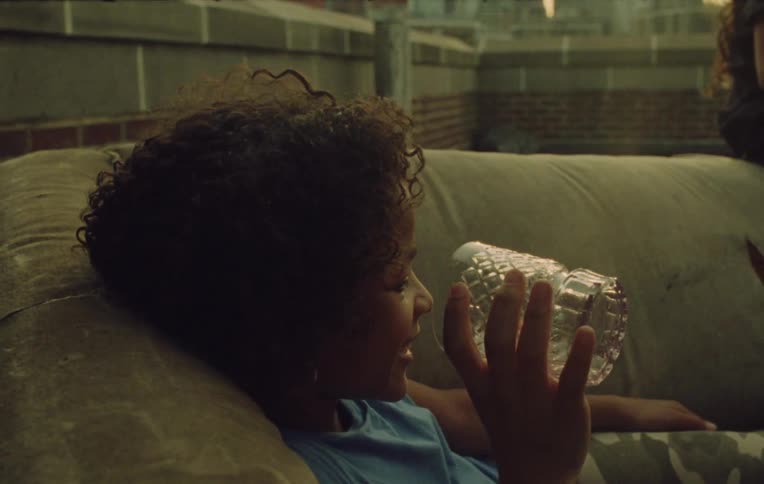 A girl lies on a couch talking to someone and inspecting a glass on a roof in a city