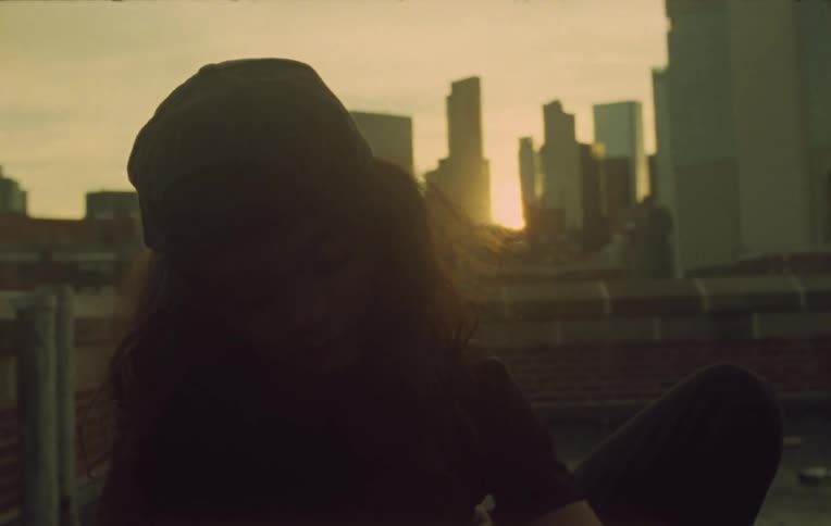 Girl wearing a baseball cap sits on the roof of a building in the city at evening