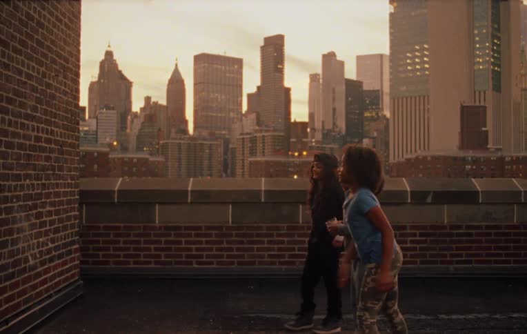 Three children on the terrace of a building facing a wall one of them gives a cup to the friend who then throws it against the wall