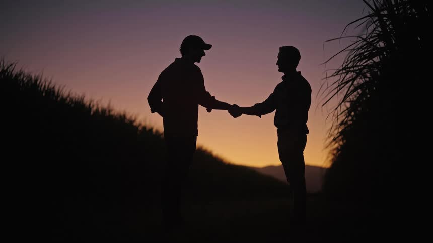 Two men shake hands on a dirt road at sunset