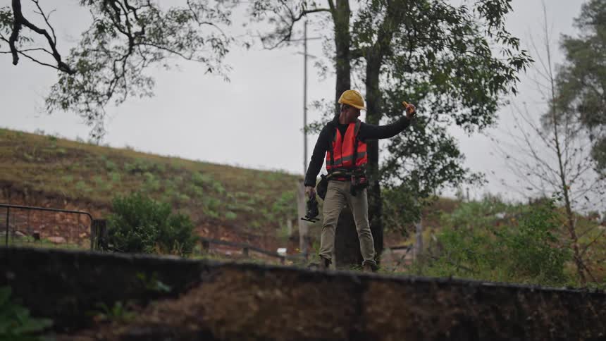 Worker wearing a vest and hardhat shines a flashlight on a ditch