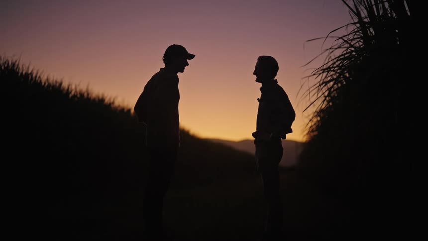 Silhouette of two men shaking hands in an outdoor agricultural setting with crops around and the sunset in the background