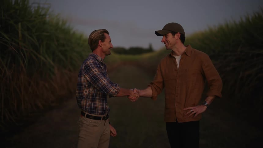 Two men laugh and shake hands standing next to fields of crops