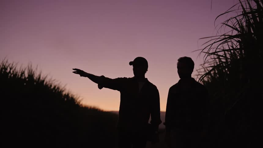 Silhouette of two men walking and pointing in some direction while conversing in the midst of an agricultural area with a reddish sky in the background