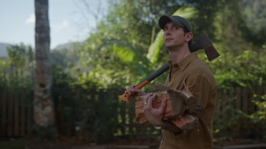 A man walking straight ahead holding an axe on his shoulder and carrying cut firewood with his other hand