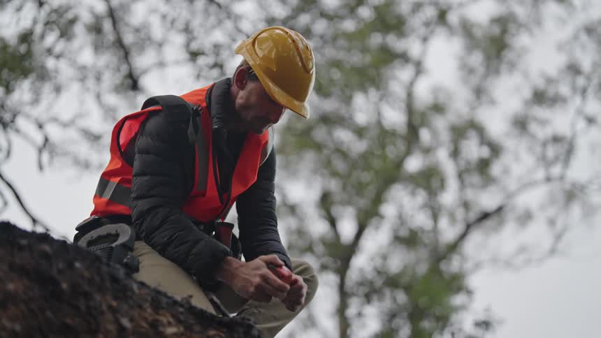 A man wearing an orange safety vest and yellow helmet kneels outside and grabs a measuring instrument out of his tool belt
