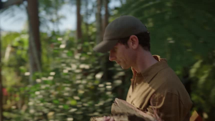 Man wearing cap walks outdoors holding wood and ax
