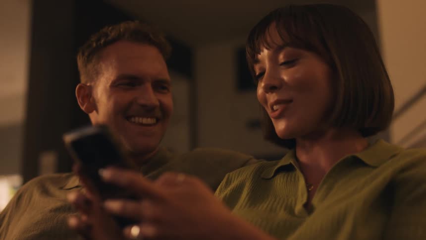 A man and woman talk and laugh sitting inside a house while she uses her phone