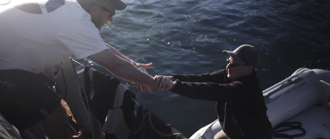 A man on a raft hands over tools to someone on board a boat