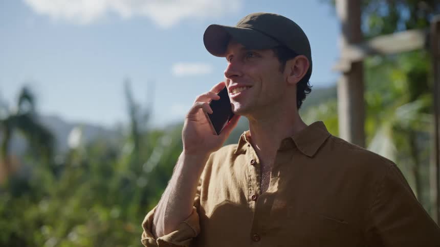 A man talks on a cell phone laughing and looks at his watch while standing outside