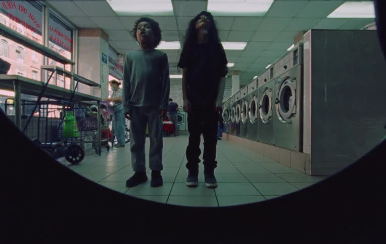 Two children standing inside a laundry establishment and then they appear in a cut of a recording scene with another child