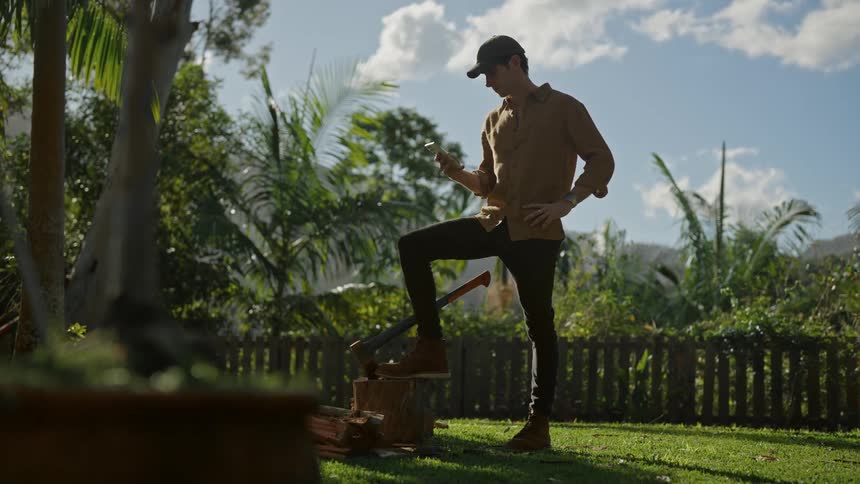 A man stands with one leg on a block of wood looking at his phone in a yard
