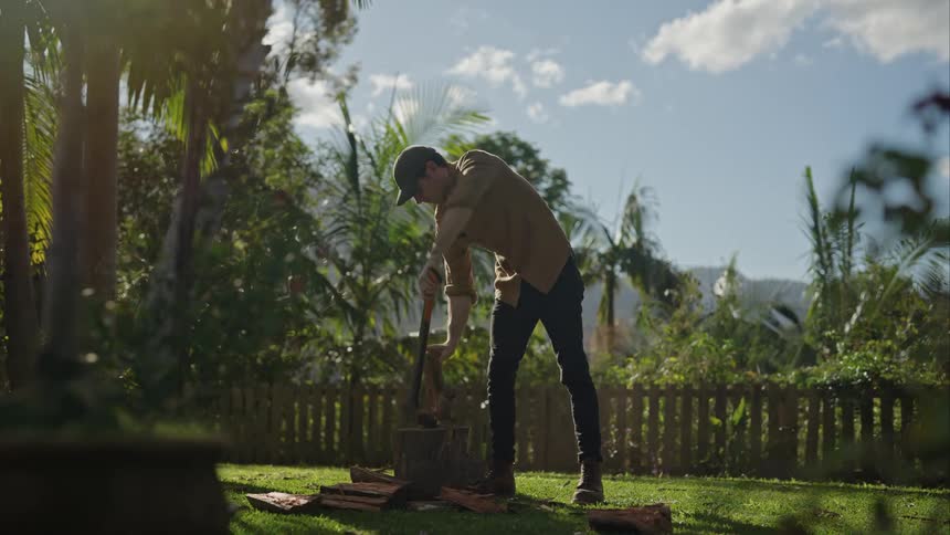 A man chops wood with an axe in a yard surrounded by trees on a sunny day