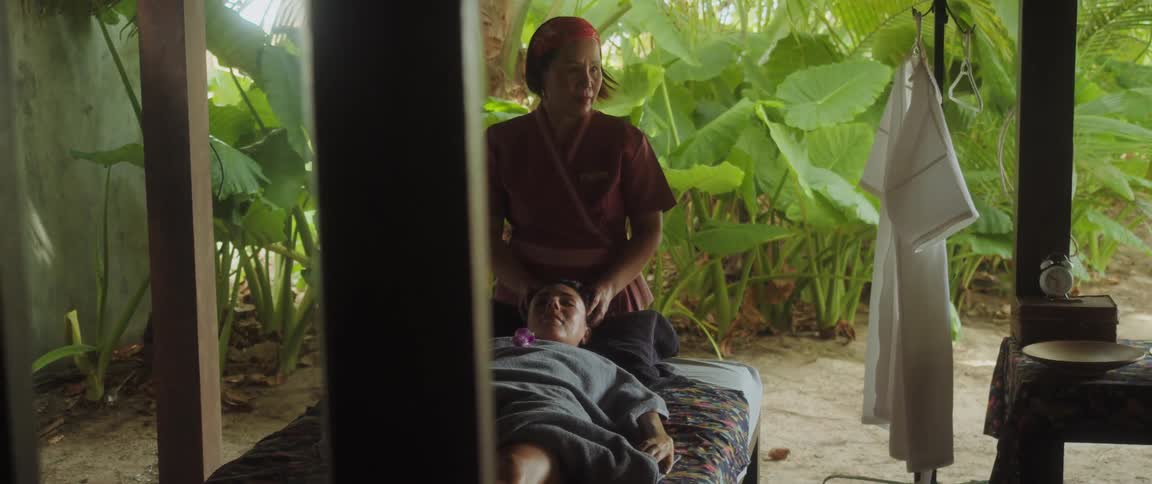 A woman massaging another woman while she lay on bed with plant in the background