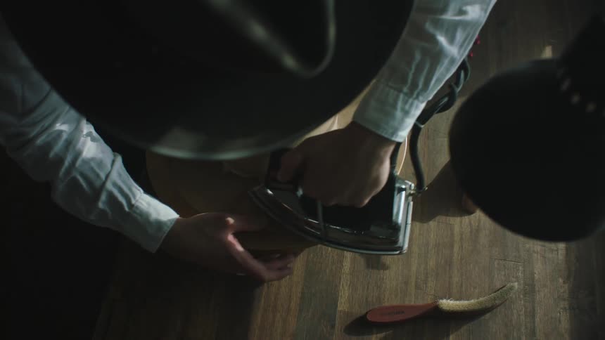 A man in a hat is hot ironing a hat that is on top of a table