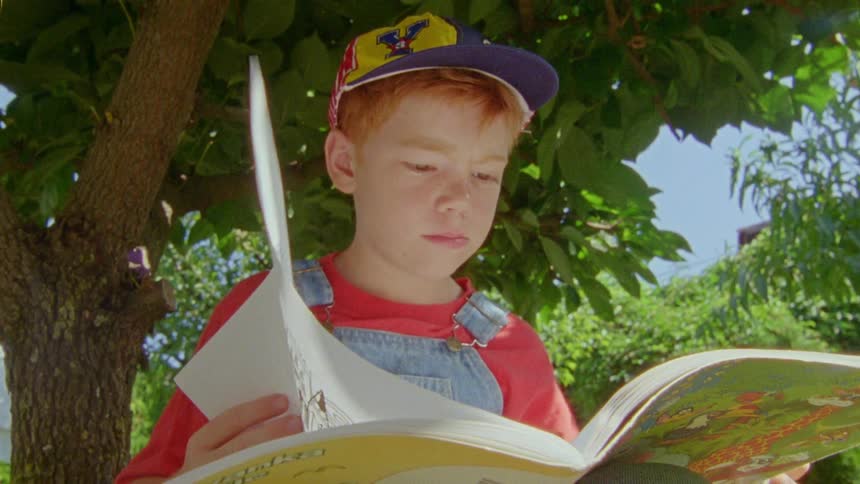 Man with cap red shirt and denim overalls reads a book in the yard