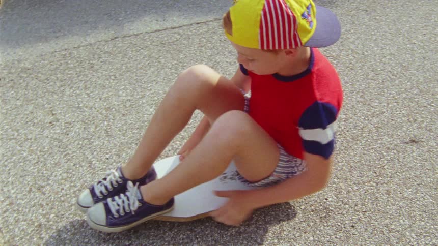 Boy with colorful cap red t shirt and shorts sits on skateboard and goes down the road
