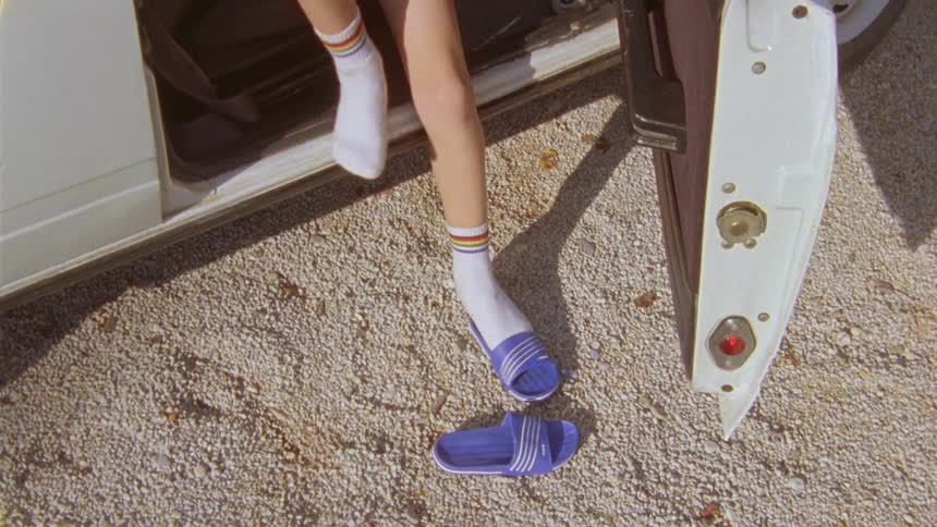 A kid opens the passenger side door of a car takes off his slippers and puts on a shoe