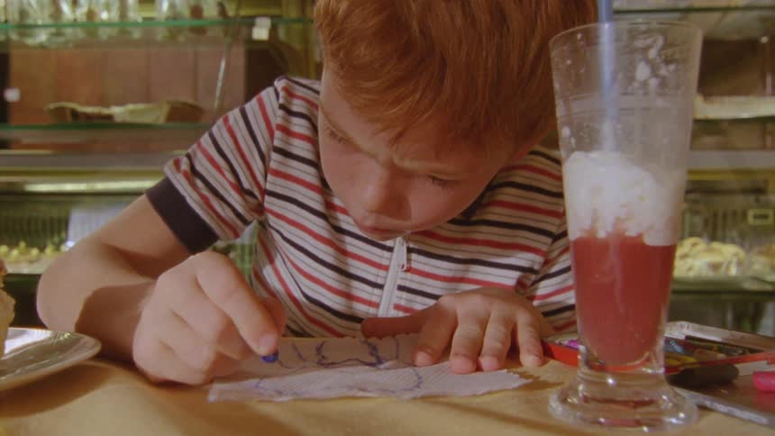 A child sits at a table in a bakery store writing on a piece of paper while a smoothie rests beside them