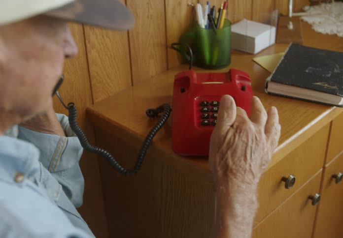 An ederly man picks up a red landline placed on a wooden shelve and place a book on top