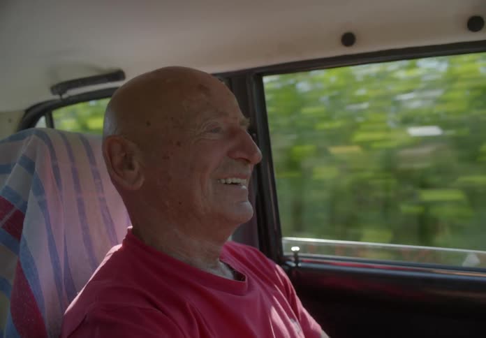 An elderly man drives a car while talking to someone and smiling