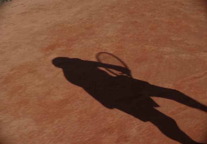 A shadow of a person on the ground walking with a tennis racket in their hand during the day