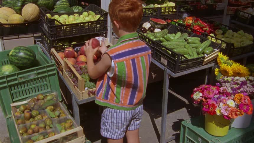 A kid walking into a fruit store picking two red apples from a basket with cash in his hands