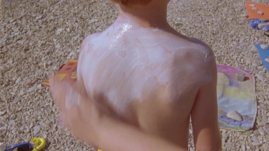 Woman applies sun cream on back of a boy on pebble beach