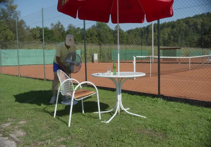 A man set up a standing fan outdoor in front of a tennis court next to a chair and a table under an umbrella