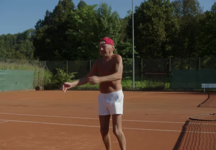 Elderly person shirtless and wearing a cap warming up and stretching his body on a tennis court