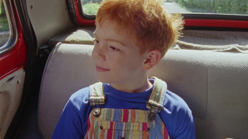 Sitting at the back of the car happy boy with red hair looks out the window